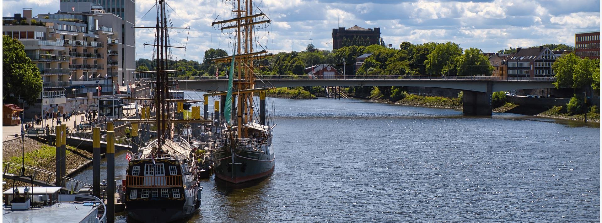 Hafenpromenade mit historischen Segelschiffen und moderner Architektur an einem Fluss unter bewölktem Himmel
