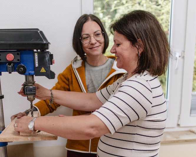 Zwei Frauen bedienen gemeinsam eine Standbohrmaschine in einem Werkraum