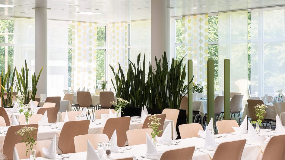 Ein heller Speisesaal mit langen Tischreihen. Im Hintergrund eine große Fensterfront und Grünbepflanzung. 