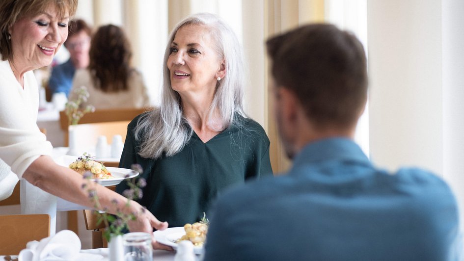 Mann und Frau sitzen im Speisesaal am Tisch und bekommen ihr Essen von einer Frau serviert
