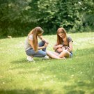 Zwei junge Frauen sitzen auf einer grünen Wiese, während eine von ihnen ein Häschen auf dem Schoß hält und die andere sich zu ihr hinüberbeugt.