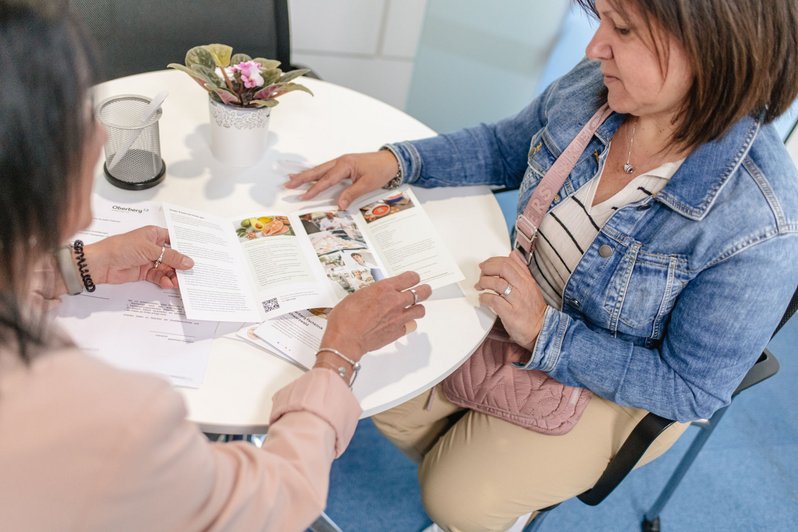 Mitarbeiterin erklärt Patientin wichtige Abläufe und zeigt in einem Flyer