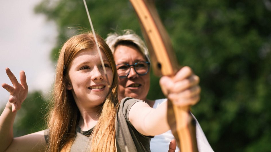 Junge Patientin mit Bogen in der Hand und Therapeutin im Hintergrund