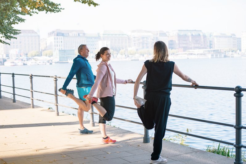 3 Personen halten sich an einem Geländer fest und dehnen ein Bein