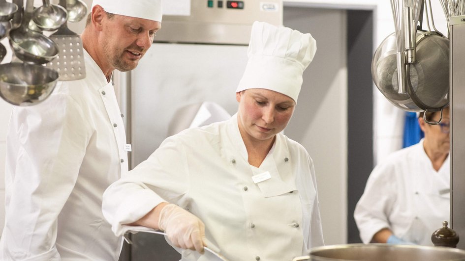 Koch und Köchen stehen in der Klinikküche. Der Koch schaut der Köchin bei Ihrem Tun über die Schulter. 