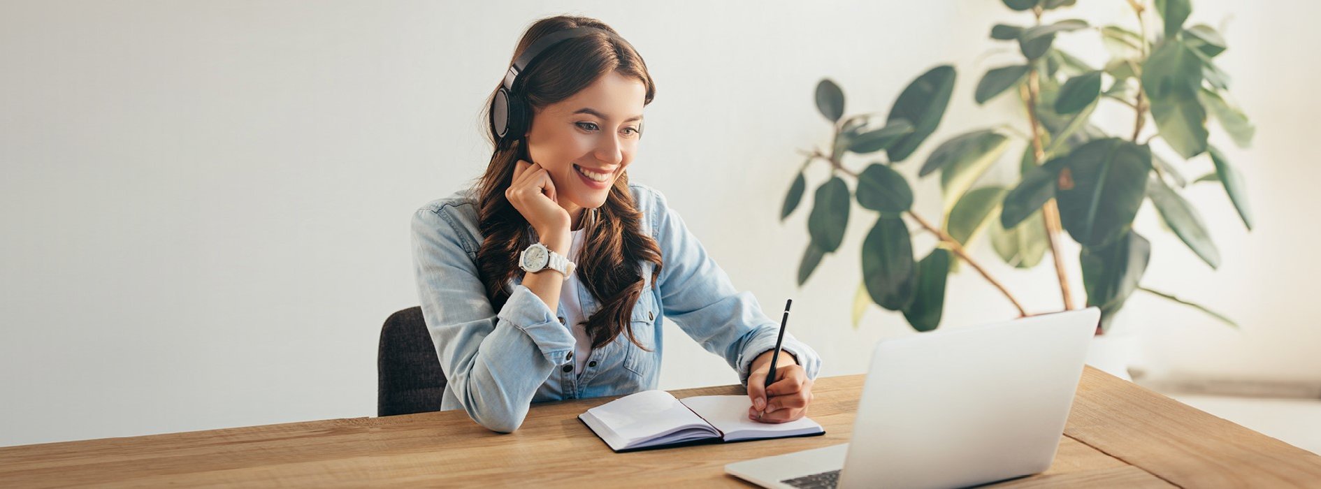 Junge Frau mit Kopfhörern sitzt an einem Schreibtisch, lächelt und macht Notizen während eines Online-Vortrags auf ihrem Laptop. Digitale Fortbildung oder Webinar in einer entspannten Umgebung.