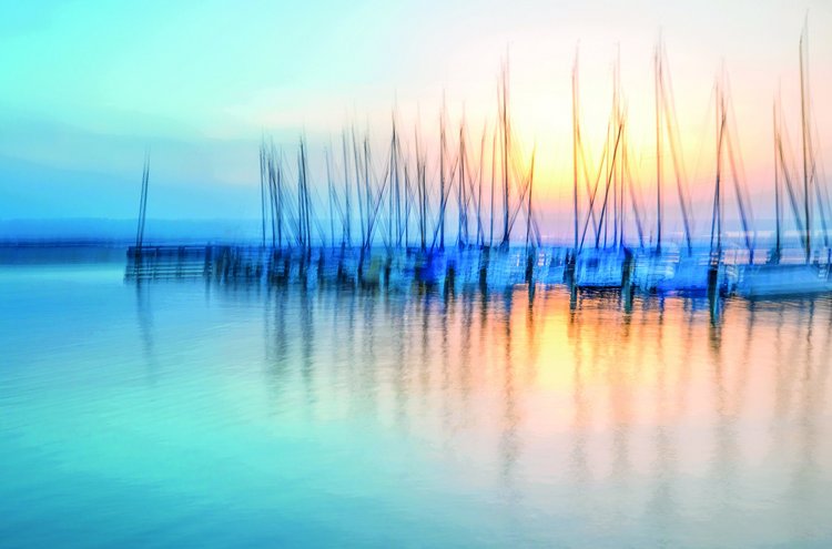 Verträumte Aufnahme eines Yachthafens im Abendlicht, mit unscharfen Bootsmasten, die sich im ruhigen Wasser spiegeln – in sanften Blau- und Goldtönen bei Sonnenuntergang