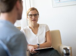 Burnout - Eine Frau im weißen Oberteil mit schwarzer Brille, die auf einer Couch sitzt mit einem Mann im Vordergrund