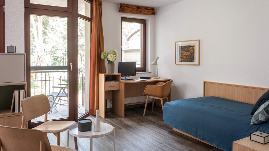 Möbliertes Patientenzimmer mit Bett, Schreibtisch, Sitzgelegenheiten und Balkon. 