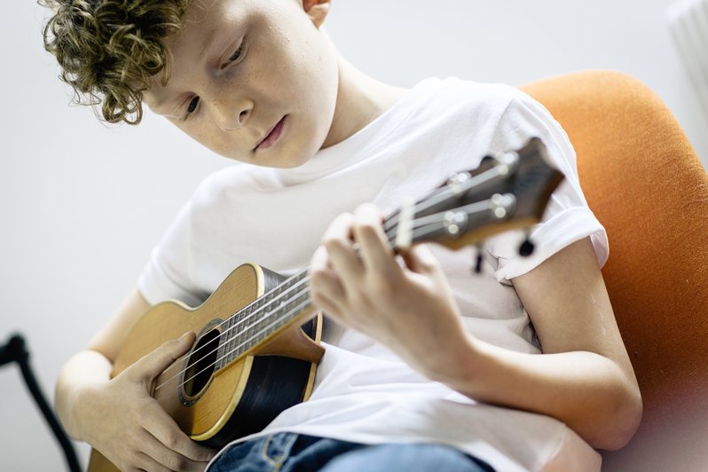 Junge sitzt auf orangenem Sessel mit einer Ukulele in der Hand und spielt
