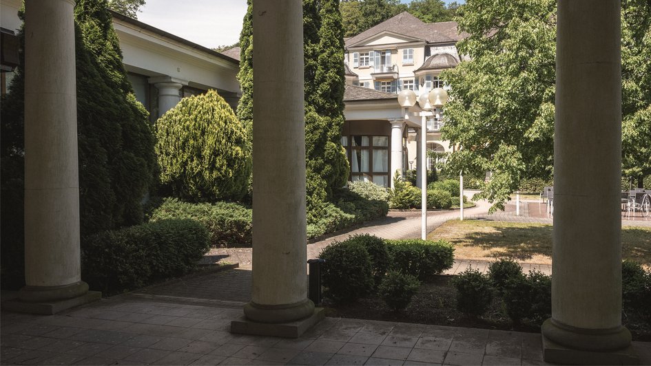 Im Vordergrund Überdachung mit Säulen, im Hintergrund hinter Bäumen versteckt ein Haus.