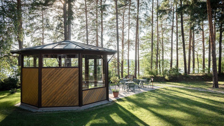 Ein Holzpavillon  auf einer Wiese. Im Hintergrund Bäume. 