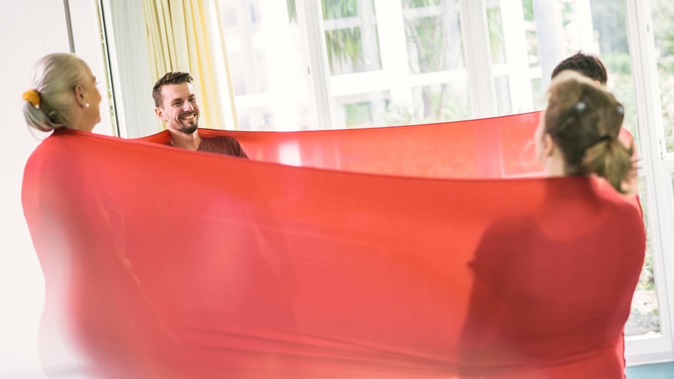 Vier Personen stehen sich gegenüber im Kreis, umwickelt von einem roten Tuch