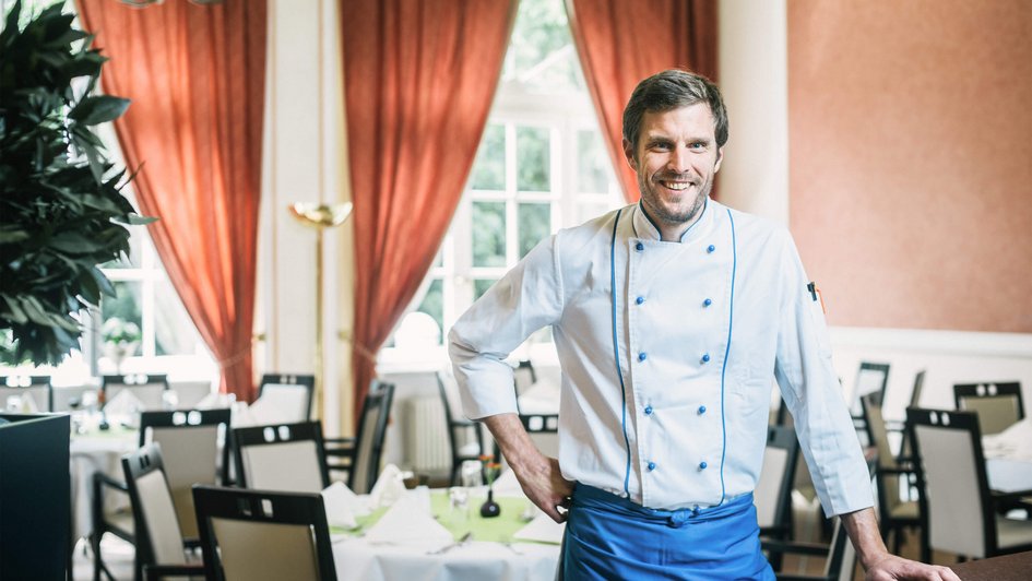 Küchenchef steht im Speisesaal, hat eine Hand in der Hüfte, die andere locker auf einem Tisch aufgelegt.
