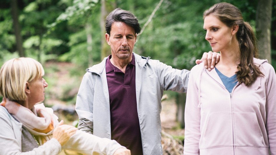 Ein Mann hat die Augen geschlossen und hat seine Hand auf der Schulter einer Frau. Eine weitere Frau beobachtet das Duo.