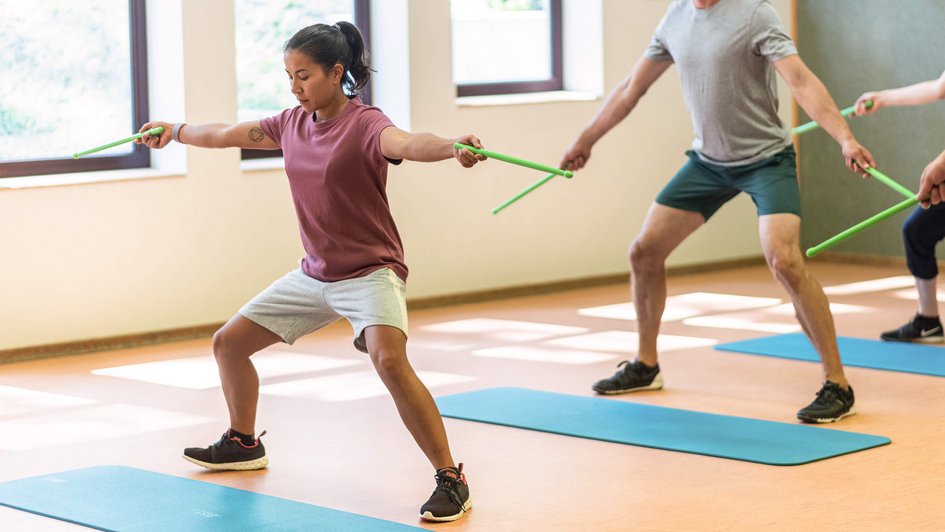 Gruppe von Menschen steht in leicht gehockter Stellung hinter einer Gymnastikmatte. Sie halten Holzstäbe in ihren ausgestreckten Händen.