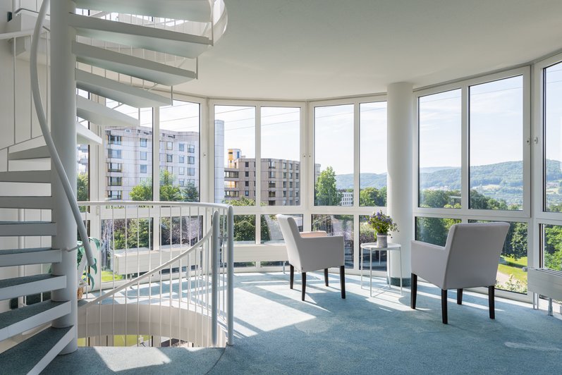 Ein lichtdurchfluteter Aufenthaltsbereich mit moderner Einrichtung, großen Fensterfronten und einer eleganten Wendeltreppe