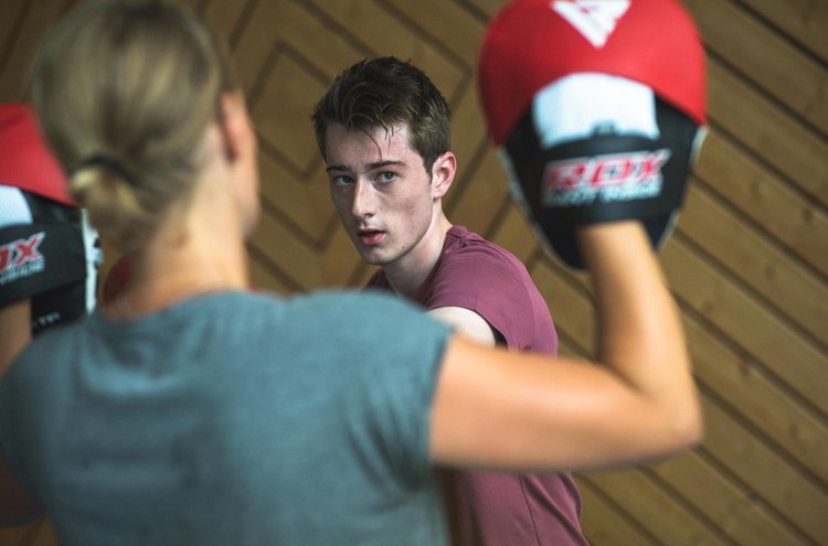 Ein junger Mann und eine Frau befinden sich in einer Sporthalle. Die Frau trägt Pad-Handschuhe, welche der junge Mann fokussiert.