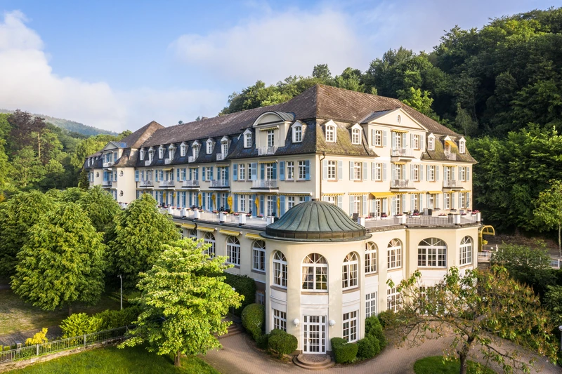Au&szlig;enansicht der Oberberg Parkklinik Wiesbaden-Schlangenbad: Denkmalgesch&uuml;tztes Geb&auml;ude vor gr&uuml;nen B&auml;umen und blauem Himmel, mit Gehweg im Vordergrund